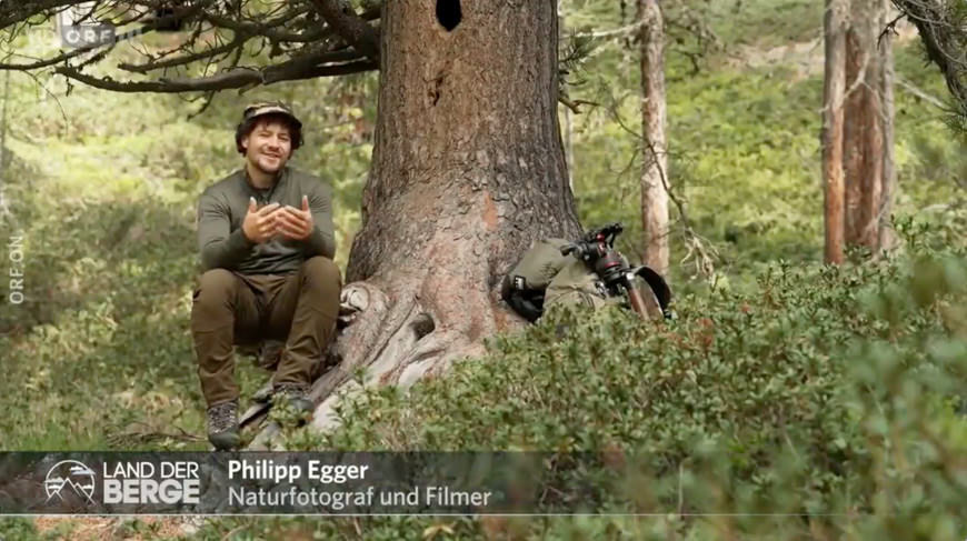 Natürlicher Naturfotograf Philipp Egger im Wald, umgeben von grünen Büschen und Bäumen, sitzt entspannt und lächelnd neben einer Kamera, die auf einem Baumstumpf liegt, während er seine Leidenschaft für die Berge und die Natur dokumentiert.