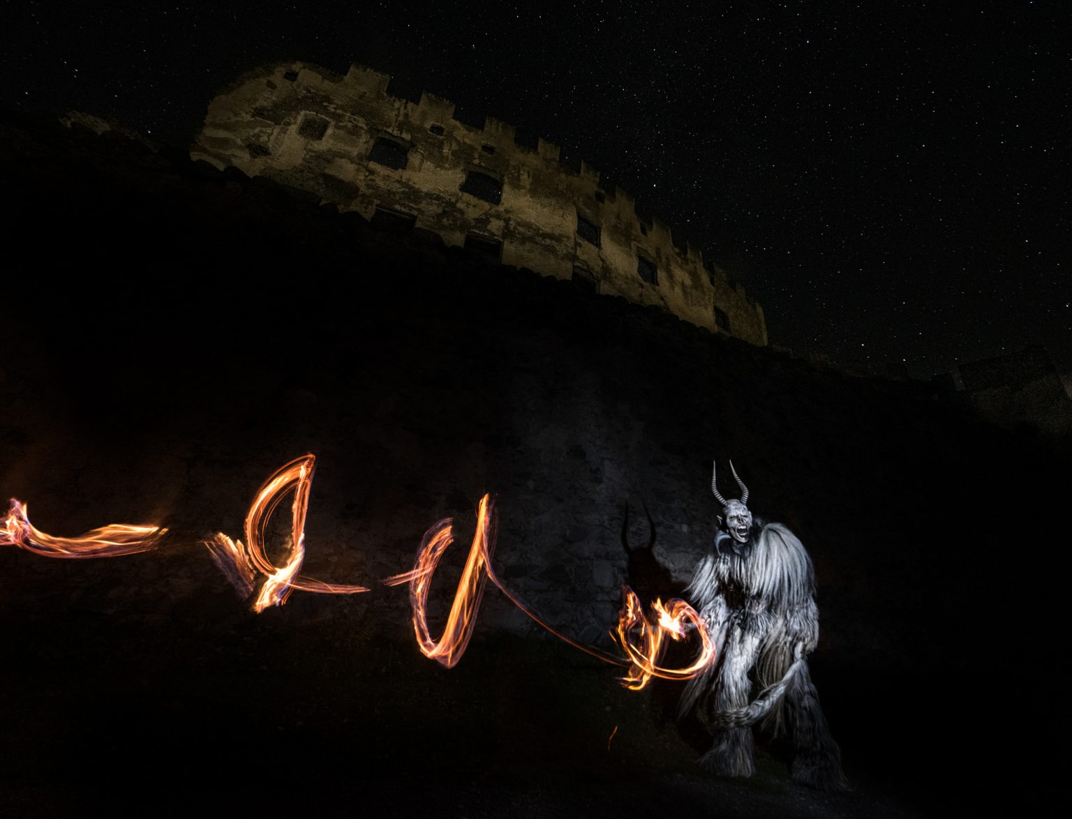 Feuriger Kostüm-Performance mit einem mystischen Wesen bei Nacht vor einer alten Burgruine, Sternenhimmel im Hintergrund für faszinierende visuelle Effekte.