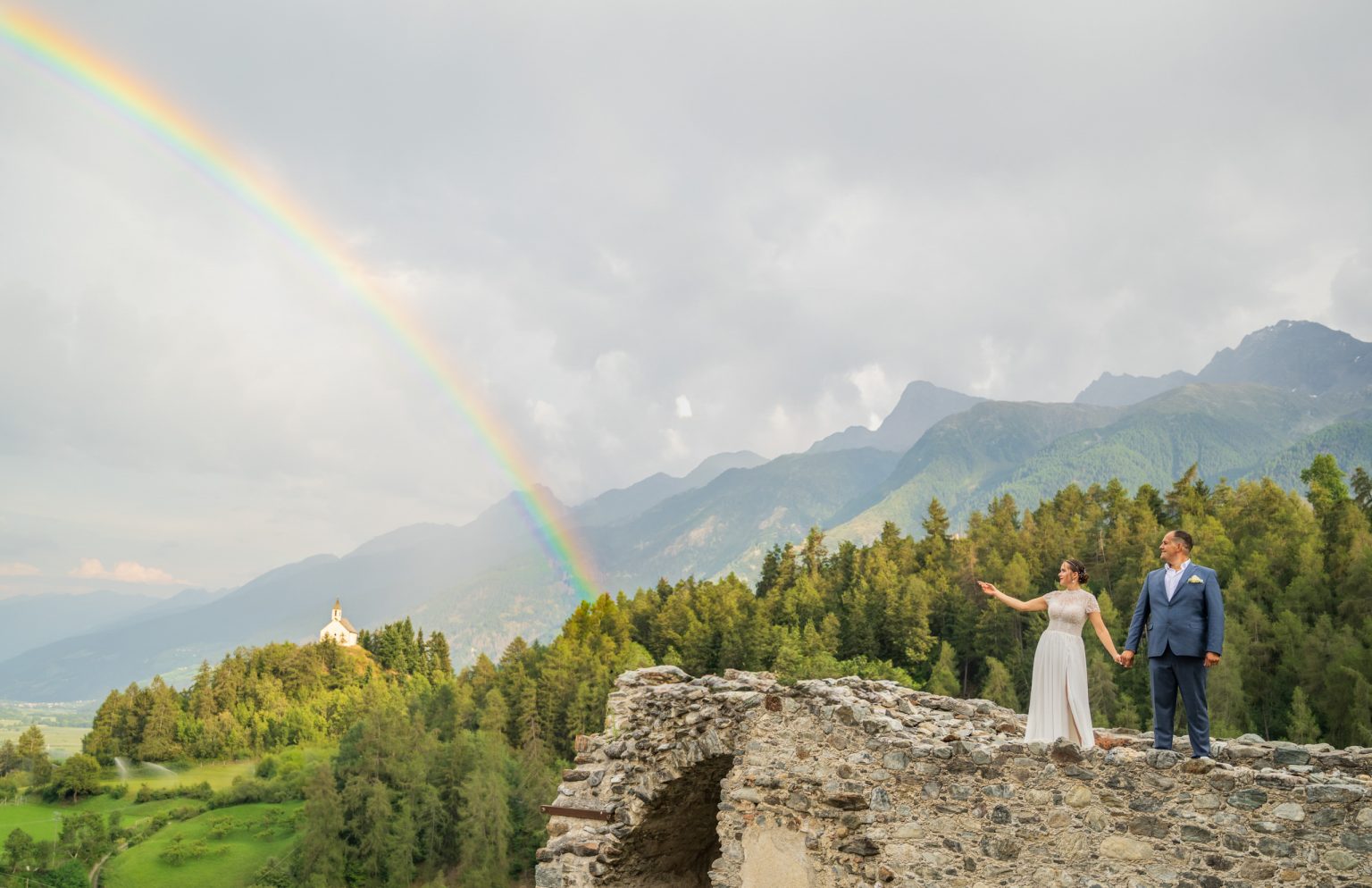 Farbenfrohe Hochzeitsfotos vor malerischer Berglandschaft mit Regenbogen, glückliches Paar in eleganten Outfits hält Händchen und genießt die Natur, perfekte Kulisse für romantische Erinnerungen.