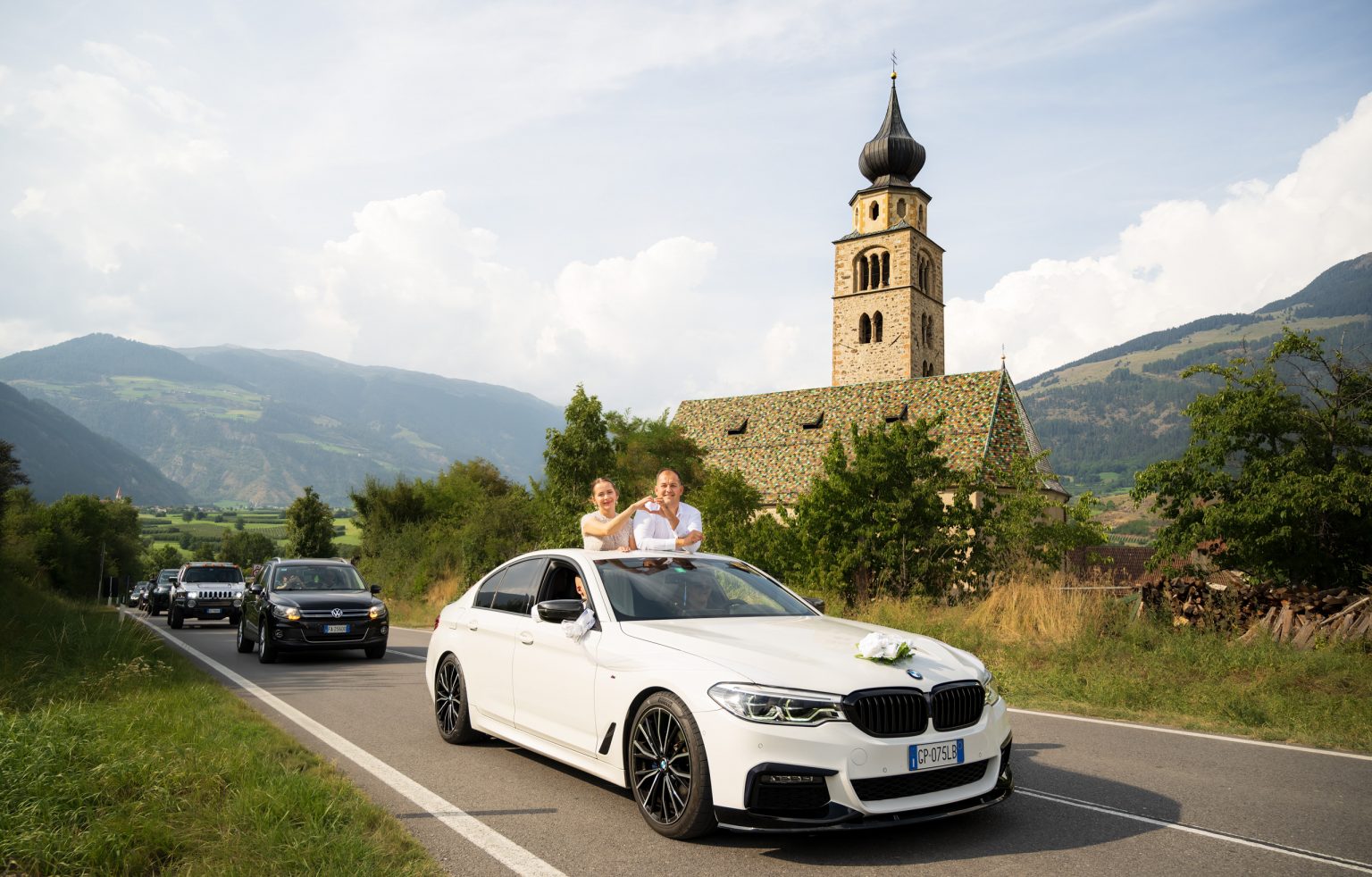 Hochzeit auf einem Landstraßen-Hintergrund mit einem weißen BMW, Paar in der Limousine, die Freude zeigt, und einer Kirche im alpinen Umfeld, bei schönem Wetter.