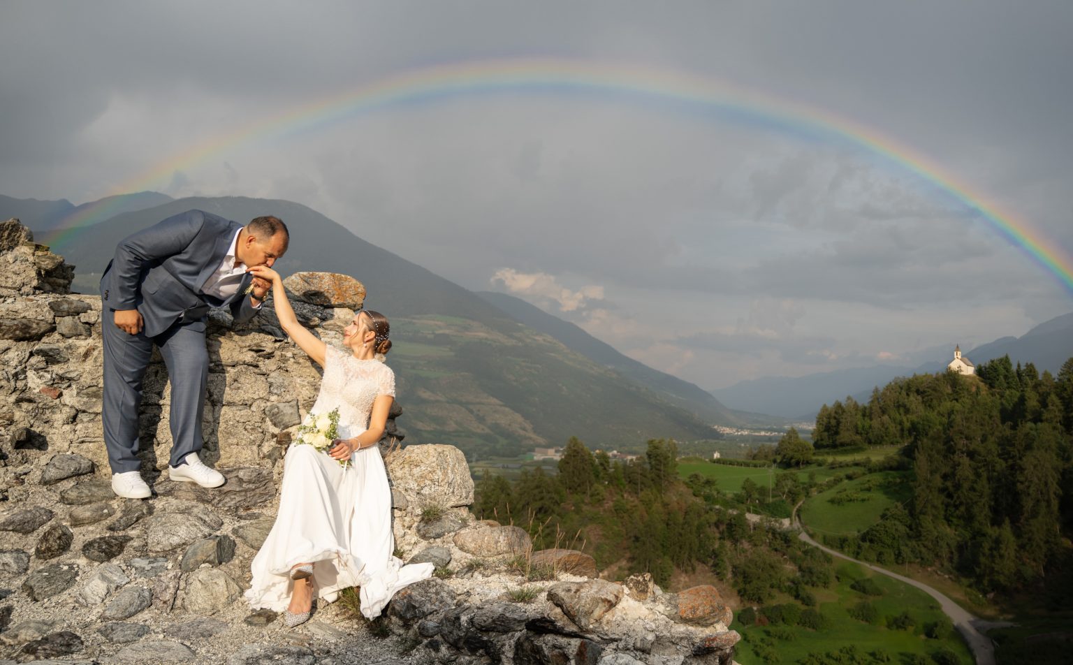 Traumhaftes Hochzeitsfoto mit Berge und Regenbogen im Hintergrund, Braut in weißem Kleid und Bräutigam im Anzug, romantische Stimmung, outdoor Naturlandschaft, besondere Hochzeitsmomente, Hochzeitsfotograf Optima.